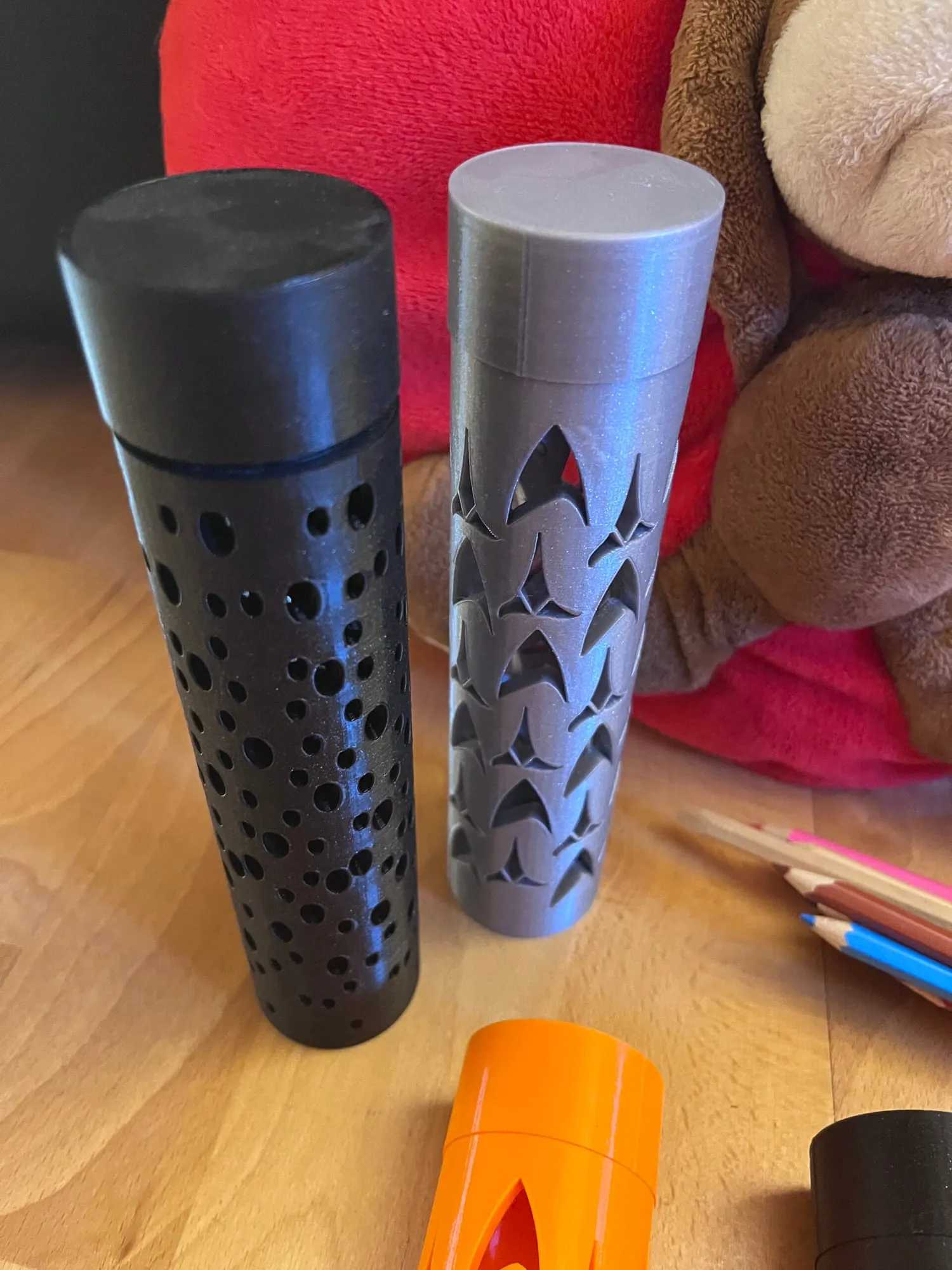 Deux étuis cylindriques ajourés, l’un noir à trous ronds, l’autre gris à motifs, près de crayons de couleur