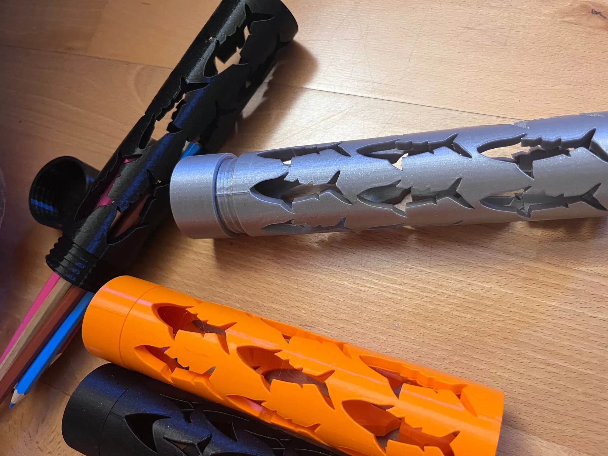 Étuis cylindriques ajourés gris, orange et noir posés sur une table en bois avec des crayons de couleur