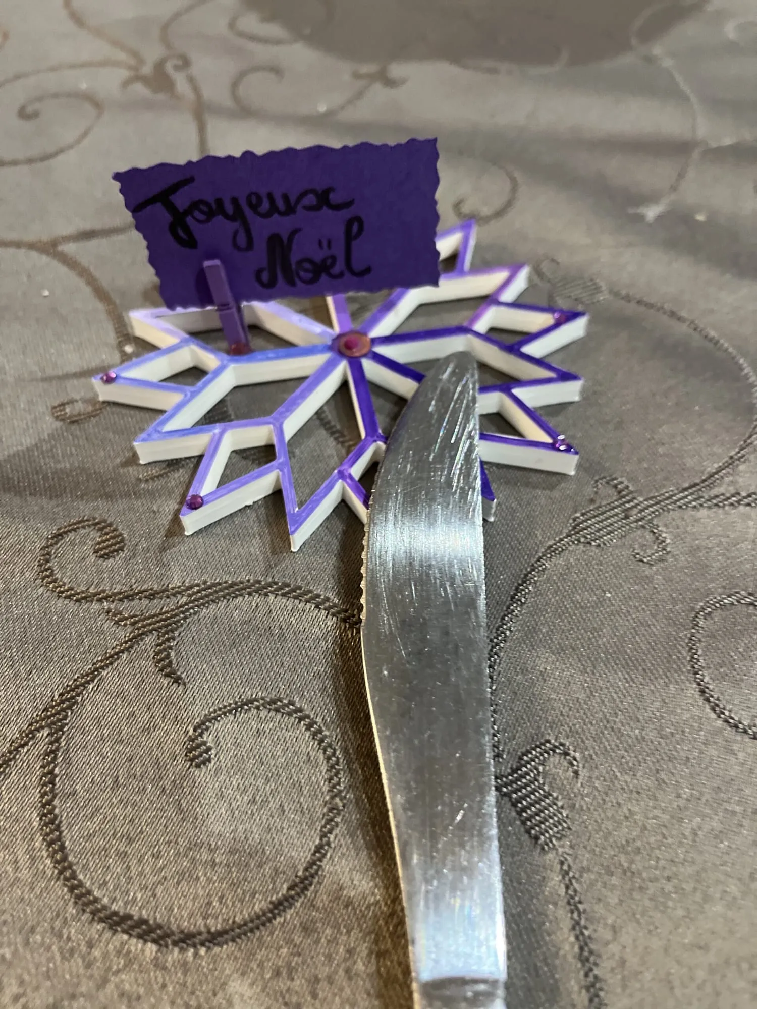 Étoile blanche décorée de violet servant de marque-place « Joyeux Noël », posée près d’un couteau