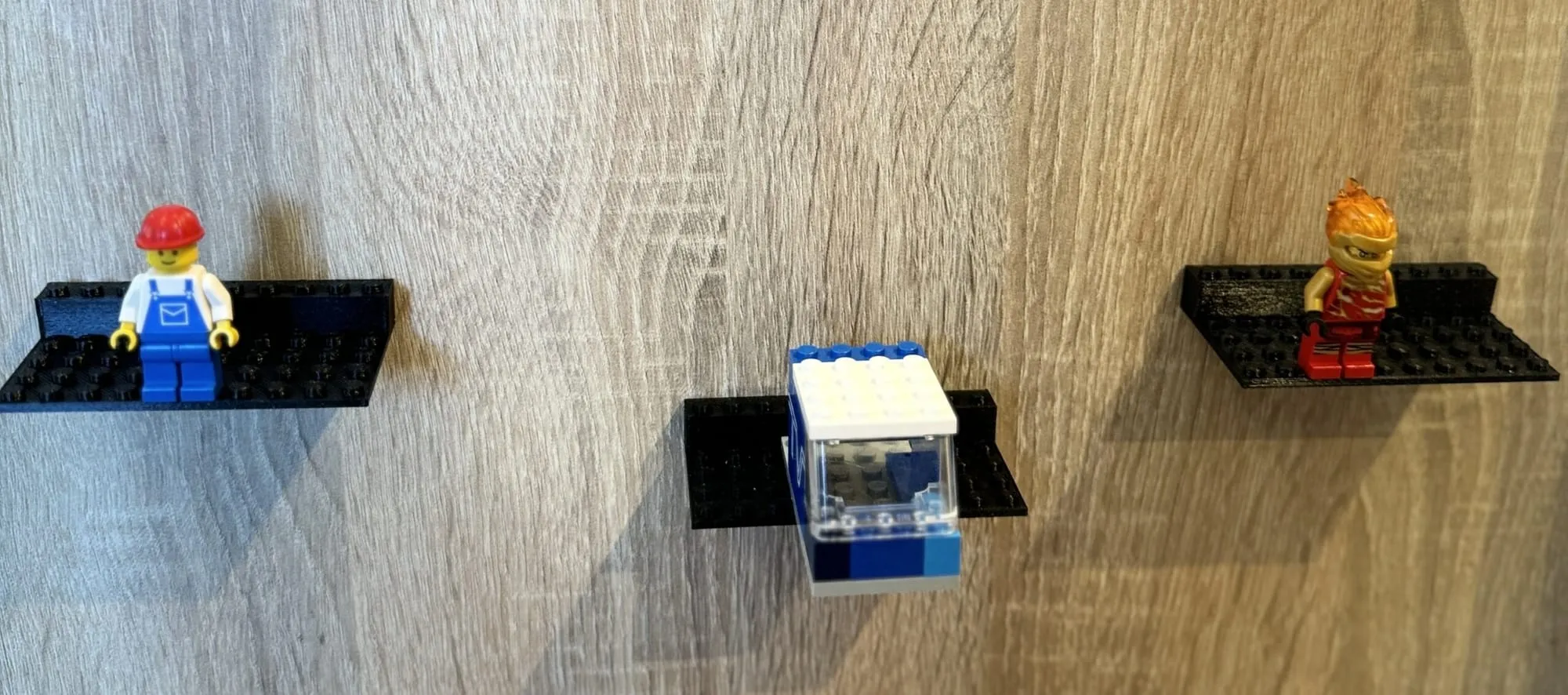 Trois supports noirs fixés sur un mur en bois, portant deux figurines et une petite construction bleue et blanche